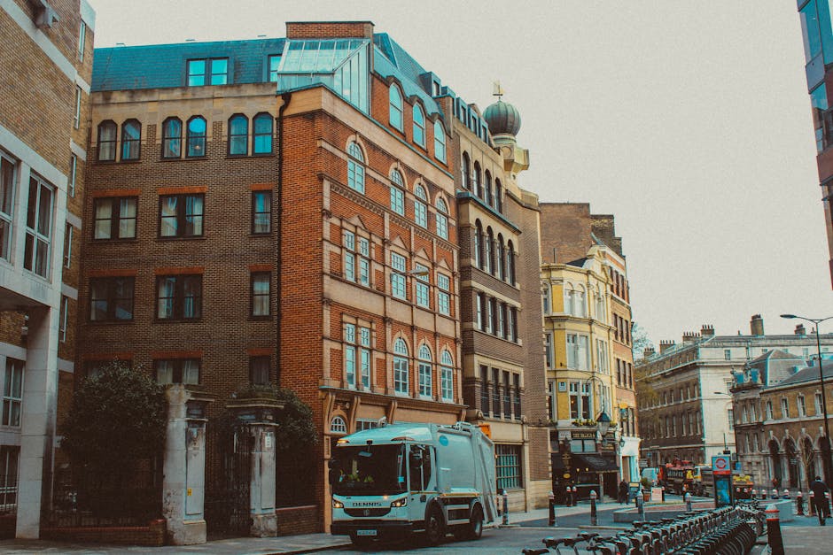 A street scene featuring a variety of multi-storey commercial and residential buildings with distinct architectural styles, including brick facades, large windows, and decorative elements. In the foreground, a light grey waste collection truck is parked on the road, partially obscuring the entrance to a gated property with stone pillars and greenery. Other vehicles and bicycles are visible along the street, which is lined with bollards and bicycle racks. The environment appears to be an urban area with a mixture of historic and modern structures, under overcast daytime lighting that provides soft, diffuse illumination. This scene relates to private waste collection services like those offered by Waste Collection Maida Vale, illustrating an everyday context for on-site rubbish removal or alternative waste handling on busy city streets, where independent waste collection providers operate alongside local authority services.