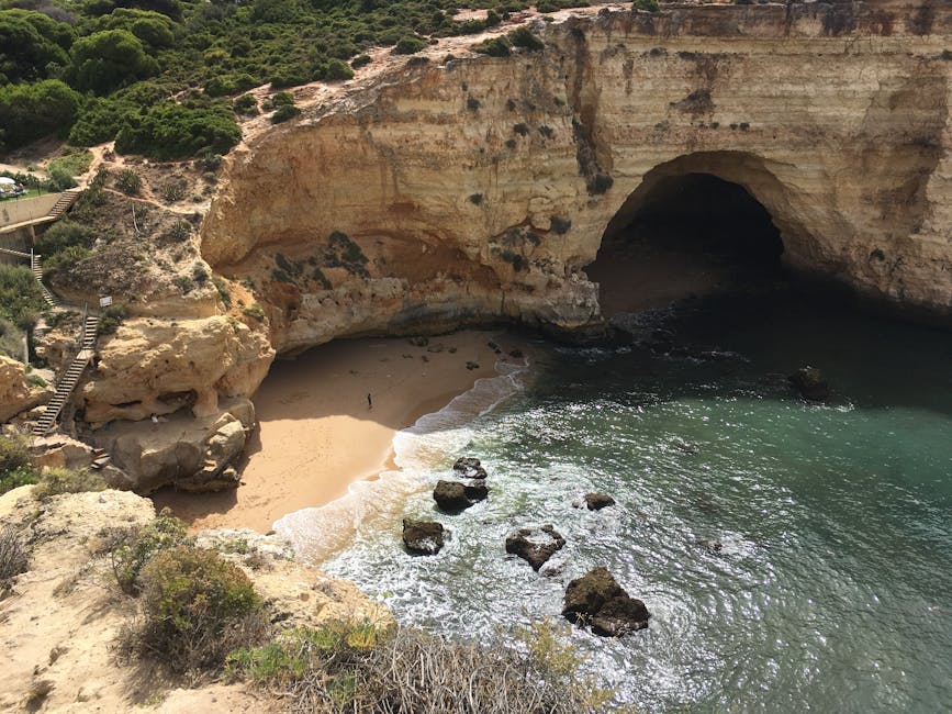 A natural coastal landscape featuring a large sea cave with a wide, dark opening in a layered limestone cliff face. The cliff exhibits shades of beige, tan, and light brown with weathered, textured surfaces and darker patches indicating natural mineral deposits. Surrounding the cave, the cliff extends to rugged terrain covered with sparse green vegetation and small bushes. To the left, a wooden staircase with railings descends along the cliffside, leading towards a small, sloping sandy beach at the water's edge. The beach has fine light-colored sand, with a few rock formations partially submerged in the gentle, rippling sea water. The ocean water displays varying tones of turquoise and deep green, with foam forming where the waves meet the shore. In the background, the coastline continues with more rocky promontories and greenery, under an overcast sky. This scene is characteristic of a secluded natural cove, often visited for private visits or alternative access for waste removal from remote coastal sites. Waste Collection Maida Vale’s services may be applicable for managing rubbish from such locations.