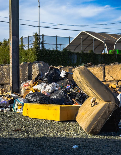 A street scene featuring a variety of multi-storey commercial and residential buildings with distinct architectural styles, including brick facades, large windows, and decorative elements. In the foreground, a light grey waste collection truck is parked on the road, partially obscuring the entrance to a gated property with stone pillars and greenery. Other vehicles and bicycles are visible along the street, which is lined with bollards and bicycle racks. The environment appears to be an urban area with a mixture of historic and modern structures, under overcast daytime lighting that provides soft, diffuse illumination. This scene relates to private waste collection services like those offered by Waste Collection Maida Vale, illustrating an everyday context for on-site rubbish removal or alternative waste handling on busy city streets, where independent waste collection providers operate alongside local authority services.