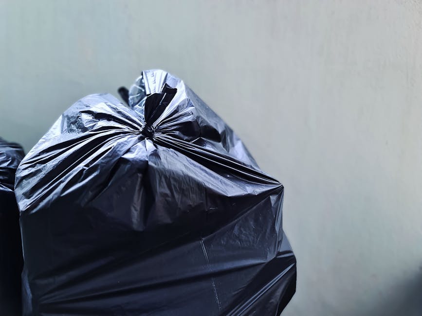 Close-up view of a black heavy-duty rubbish bag made from glossy plastic material, tightly knotted at the top, resting against a plain, light-colored wall. The bag appears partially filled, with visible folds, creases, and some minor wrinkles on its surface. The surrounding environment suggests an indoor or enclosed space, with no visible background elements other than the neutral wall. The lighting is soft, evenly illuminating the bag and highlighting its shiny texture. The scene relates to waste management and rubbish removal, consistent with services offered by Waste Collection Maida Vale, as the bag exemplifies typical refuse prepared for collection or disposal beneath the relevant local regulations. The image emphasizes an aspect of private waste handling, such as on-site clearance or independent rubbish collection, which aligns with the context of alternative refuse disposal methods.
