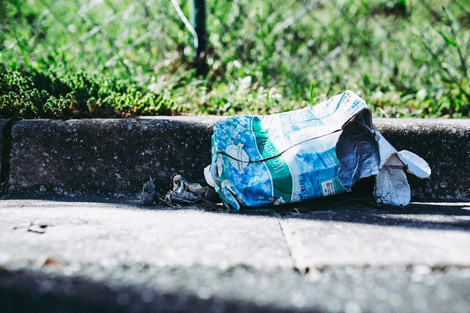 A crumpled plastic bag with blue, green, and white colors and printed text lies discarded on a concrete pavement near a weathered curb. The bag appears to be made of thin, flexible plastic material and is partially open, revealing some dark debris or waste inside. Surrounding the bag, the pavement shows signs of dirt and minor cracks. In the background, out of focus, there is a patch of lush green grass and small plants, indicating the scene is outdoors, possibly in a residential or urban area. The lighting highlights the textures of the plastic and concrete, emphasizing the environmental impact of littering. This scene reflects the importance of responsible waste disposal and the benefits of professional rubbish removal services, such as those provided by Waste Collection Maida Vale, for maintaining clean and sustainable environments in urban settings.
