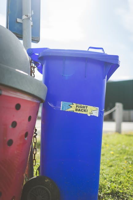 A tall, upright blue plastic waste bin with a closed lid, positioned outdoors on a grassy area near a pavement. A label with the words 'FIGHT BACK!' is taped across the front of the bin. To the left, part of a red and black wheeled rubbish container with a textured surface and a lid is visible, leaning slightly against a metal post with chains hanging from it. In the background, a partly cloudy sky and a section of a road or driveway with a metal barrier are seen, indicating an outdoor rubbish collection setting. The scene suggests an environment for private or alternative waste handling, highlighting the use of individual waste containers for rubbish disposal or collection services, such as those provided by Waste Collection Maida Vale.