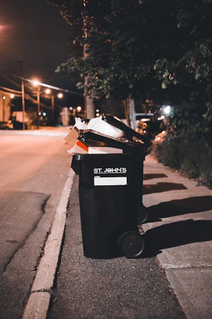 A black wheelie bin labeled 'St. John's' is positioned on a sidewalk at night, with its lid open and filled with various types of rubbish, including cardboard and plastic waste. The bin is situated near a curb on an urban street, with streetlights illuminating the scene and casting shadows on the pavement. In the background, there are trees with dense foliage on the right side, and some parked cars and lamp posts further down the street, creating a quiet, residential atmosphere. The scene appears to depict a private waste disposal setting, possibly indicating an alternative or independent rubbish collection service, with the bin placed for collection or disposal outside a property. The environment is well-lit, with the night sky slightly visible through the trees, emphasizing the urban context and the importance of effective rubbish removal services provided by Waste Collection Maida Vale.