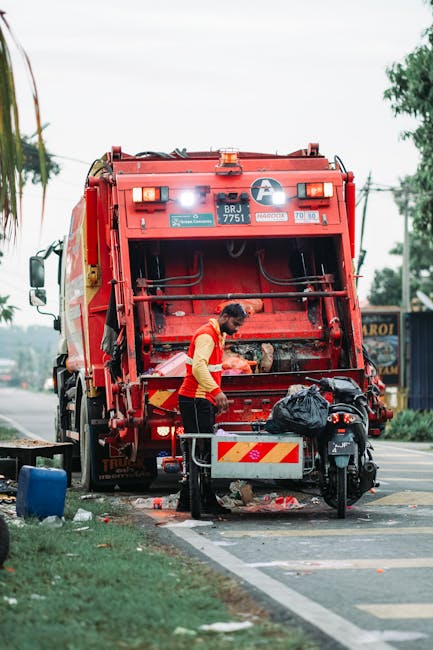 A municipal waste collection worker in a red and yellow uniform is standing at the rear of a large, red refuse collection truck, which is positioned on the side of a suburban street. The truck's rear loading mechanism is open, revealing a large, open compartment with a textured metal surface and hydraulic components. The worker appears to be handling waste or preparing the vehicle for collection, with a black garbage bag placed on a small cart in front of the truck. The scene occurs during daylight, with overcast skies providing diffuse lighting, and in the background, there are trees, utility poles, and a shop with signage visible along the roadside. The truck is parked partly on the curb, close to a designated waste collection zone, where curb side waste is visible on the ground, indicating a street-based rubbish collection point. The environment suggests an organized urban or suburban setting, typical for routine waste disposal services, and incidental to the context of alternative rubbish removal methods, it exemplifies standard municipal waste collection operations in a residential area.