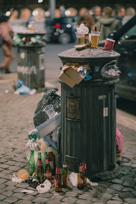 A pile of mixed waste is accumulated at the base of a brick wall with a concrete block section, located outdoors on a dirt surface. The waste includes several flattened cardboard boxes of varying sizes and a large, textured white sack, all partially open and showing signs of recent disposal with crumpled and torn edges. Some cardboard boxes reveal their corrugated material and printed labels, while others are plain brown. A blue and white plastic bag featuring a logo or branding is also visible among the debris. The arrangement suggests an informal collection of household or commercial waste, potentially prepared for removal by a private waste collection service such as Waste Collection Maida Vale. The background features a rough, weathered brick wall and a dark tree trunk to the left, with natural lighting casting shadows on the scene, emphasizing the texture of the materials and environment, illustrating a typical on-site rubbish accumulation before collection or disposal.