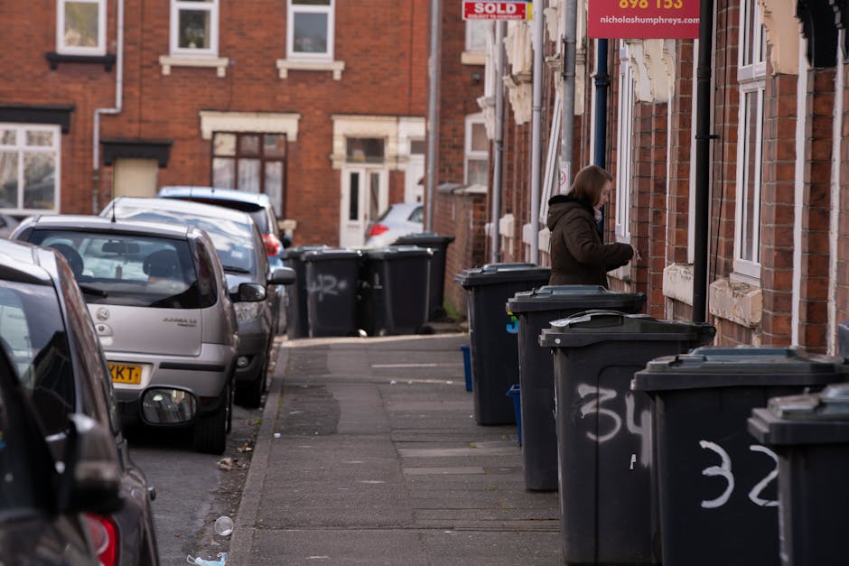 A row of black wheelie bins lined up along the pavement outside a brick residential building, with some bins partially open revealing mixed waste inside. The bins are positioned close to the front wall of the building, which features white-framed windows and stone sills. A woman wearing a dark jacket stands nearby, appearing to be managing the bins or waste disposal. In the background, additional bins are aligned along the same street, with parked cars occupying the roadway, including a silver car in the foreground. The scene is set on a typical urban street, with a mix of parked vehicles and bin storage for domestic waste collection. The environment is illuminated by natural daylight, with some litter visible on the pavement. This scene demonstrates an example of private waste handling, possibly arranged through a local waste collection service specializing in same-day rubbish removal for residential properties.
