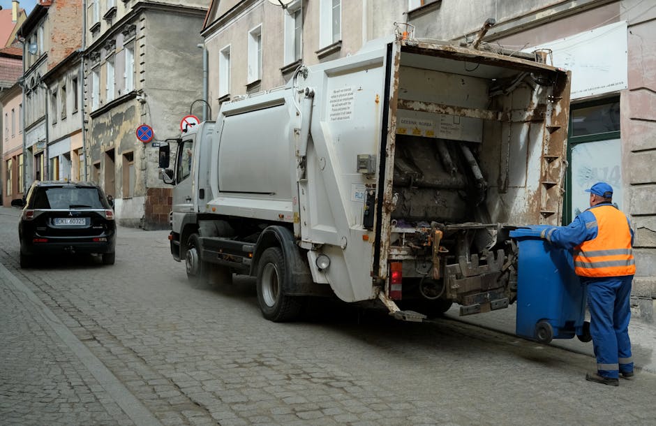 A street scene showing a waste collection vehicle parked alongside a row of older, multi-storey buildings with weathered facades, some featuring visible urban wear and tear. The white refuse truck has an open rear compactor area, revealing mechanical components and rusted metal surfaces, indicating it is used for rubbish collection and disposal. A worker dressed in a high-visibility orange vest, blue uniform, and a matching blue cap stands on the right, aligning a blue wheeled bin toward the truck's loading mechanism. The street is paved with cobblestones, and a black car is parked further along the curb on the left side. In the background, there are muted tones of grey and beige building exteriors, with a few traffic signs attached to the wall, ensuring compliance with urban waste management regulations. The scene is lit with natural daylight, emphasizing the industrial and functional environment associated with private waste removal services, reflecting the activity of independent rubbish collection firms like Waste Collection Maida Vale.