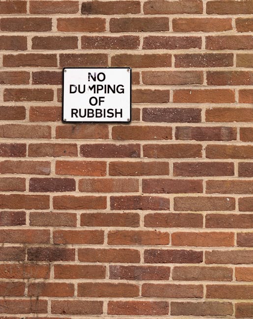 A rectangular black and white metal sign mounted on a red brick wall, displaying the message 'NO DUMPING OF RUBBISH' in bold black letters, with some of the text partially obscured or faded. The brick wall has a uniform pattern of reddish-brown bricks with light-colored mortar joints, extending across the background. The sign is positioned towards the upper left area of the image, with the wall occupying the remaining space, suggesting an outdoor setting related to waste management or rubbish disposal regulations. The overall scene appears well-lit with natural light, highlighting the texture of the bricks and the sign's surface, subtly indicating signage for private waste control or alternative rubbish removal at this location.