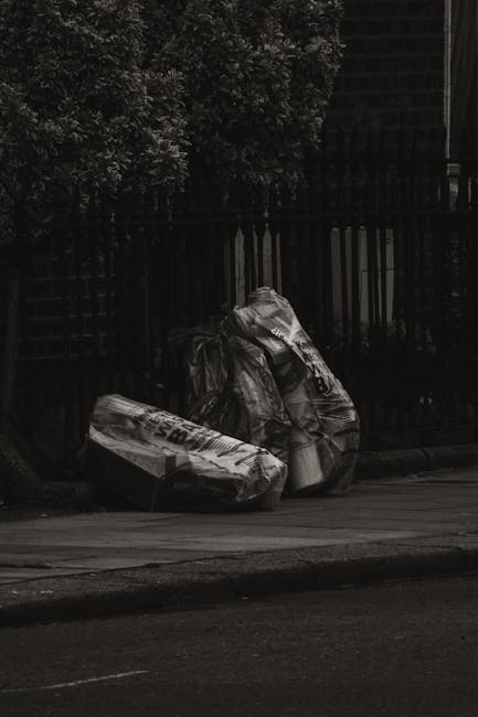 Three large black garbage bags made of thick plastic material are placed on the pavement beside a curb, leaning slightly against a black metal fence with vertical bars. The bags appear to be filled with household waste and are tightly twisted at the top. Behind the fence, there are the lower parts of dense foliage from trees or bushes, creating a natural backdrop. The scene is captured during the daytime with a natural light source, highlighting the glossy texture of the plastic bags and the matte finish of the pavement. To the right, part of a building with horizontal siding is partially visible in the background. The placement of the bags on the pavement suggests an instance of private waste disposal, potentially handled by a local rubbish removal service such as Waste Collection Maida Vale, reflecting the broader context of on-site or independent rubbish collection typically associated with non-local authority waste management.