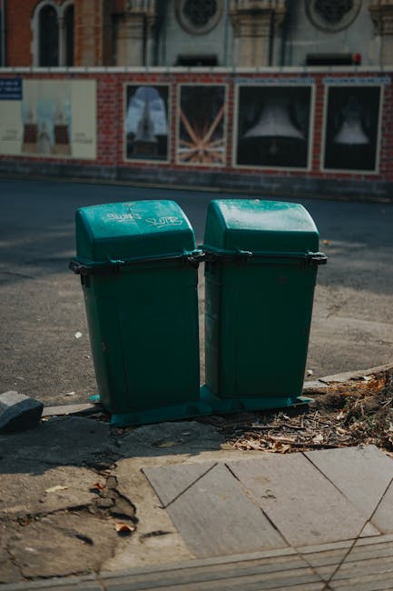 Two green wheeled wheelie bins with domed lids positioned side by side on a paved sidewalk edge beside a curb, with a small pile of dried leaves and dirt accumulated at their base. The bins display minimal branding in white lettering on the lids, indicating they are for waste disposal. Behind the bins, an asphalt road is visible, with a brick building facade featuring decorative arched windows and painted murals or advertisements. The scene is illuminated by natural daylight, casting soft shadows, and appears to be part of an urban environment where private waste collection services, such as those provided by Waste Collection Maida Vale, may be employed. The sidewalk has a mixture of concrete paving slabs and uneven, worn paving, with some small cracks and dirt, highlighting typical street-level waste management infrastructure used for rubbish disposal and collection in residential or commercial areas.